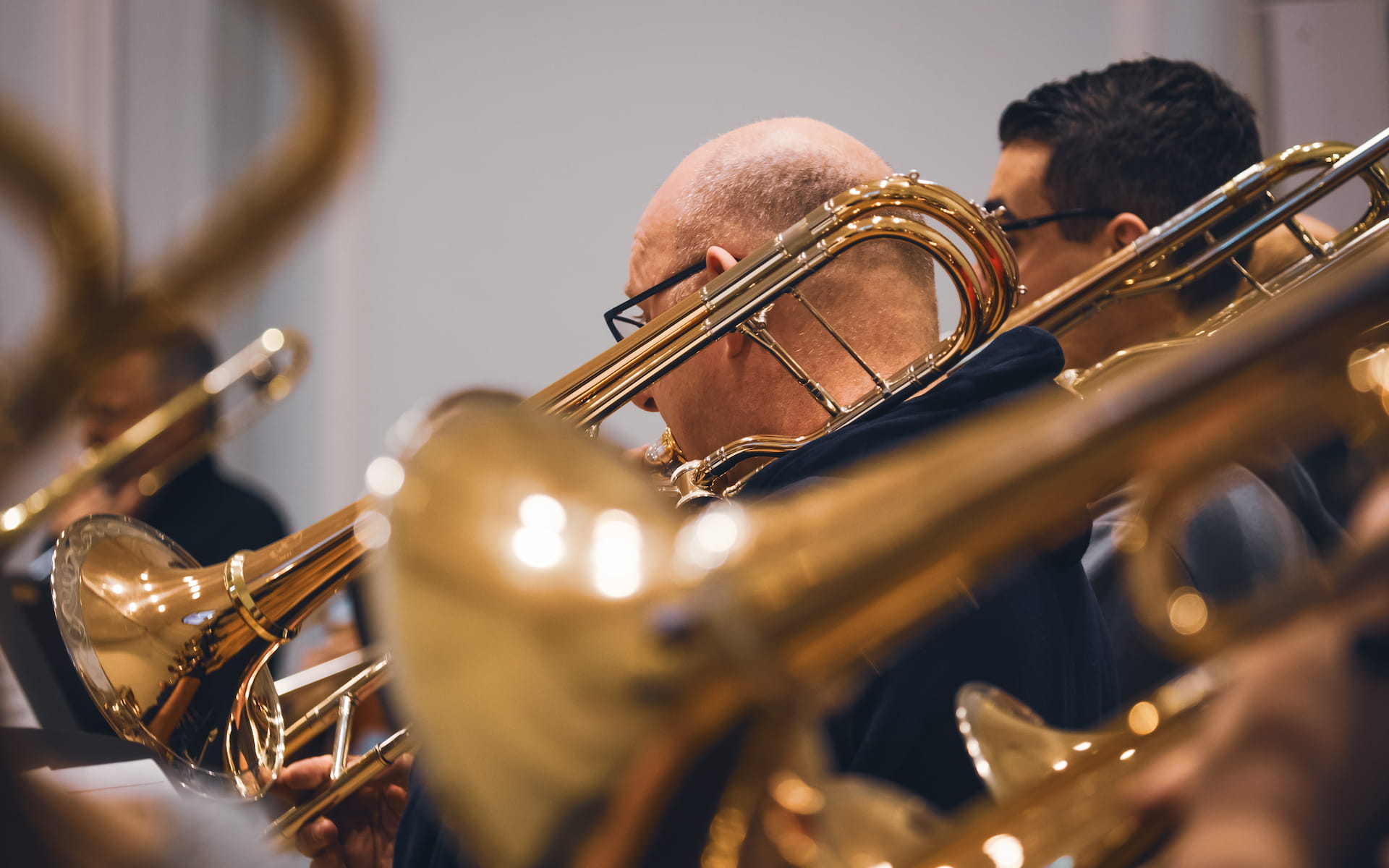 Ein Blasorchester während einer Probe: Im Fokus steht ein älterer Mann mit Brille, der eine goldglänzende Posaune spielt. Im Hintergrund sind weitere Musiker mit ähnlichen Instrumenten zu sehen, leicht unscharf dargestellt. Die Reflexionen auf den Messingoberflächen erzeugen eine warme, lebendige Atmosphäre.