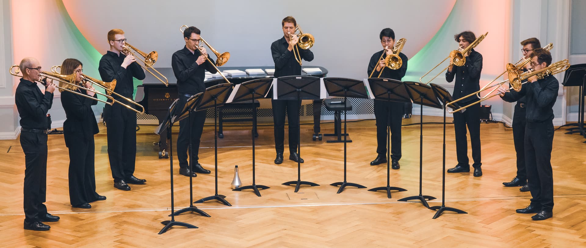 Posaunen-Ensemble in Konzertsaal: Zehn Musiker und Musikerinnen in schwarzer Kleidung spielen Posaunen im Halbkreis vor Notenständern. Klassische Konzertatmosphäre mit stilvollem Holzfußboden und dezent beleuchtetem Hintergrund.