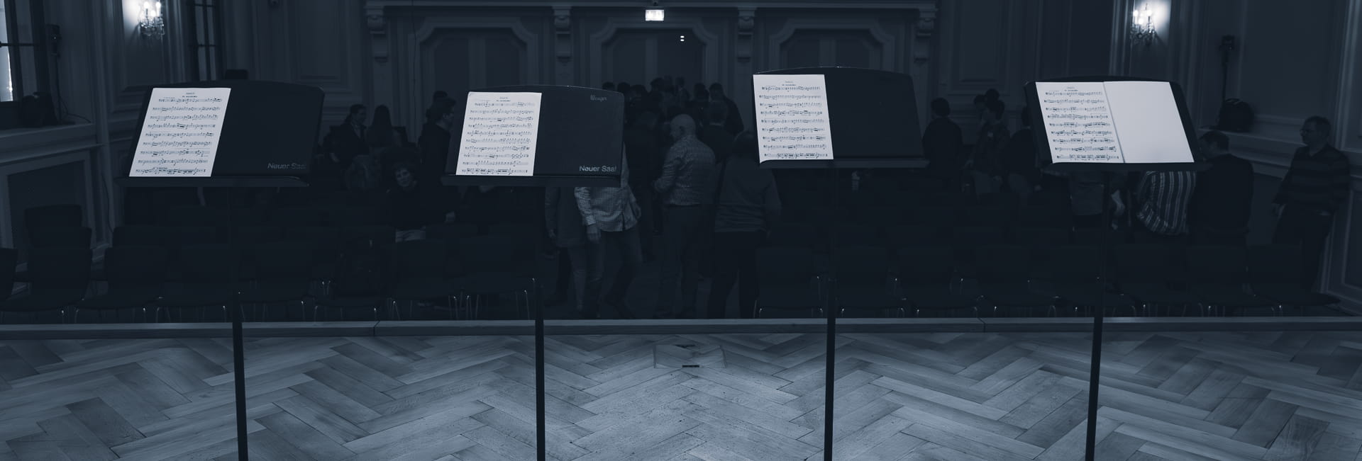 Notenständer mit Partituren in einem Konzertsaal. Vier Notenständer stehen auf einer Holz-Bühne, jeweils mit offenen Notenblättern. Im Hintergrund sind unscharf Musiker und Publikum in einem klassisch eingerichteten Konzertsaal mit Stuckverzierungen und Wandleuchten zu sehen.